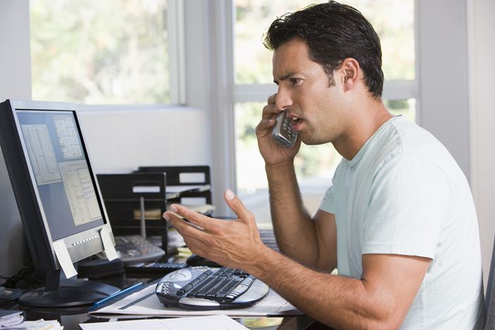 Man in home office on telephone using computer and frowning